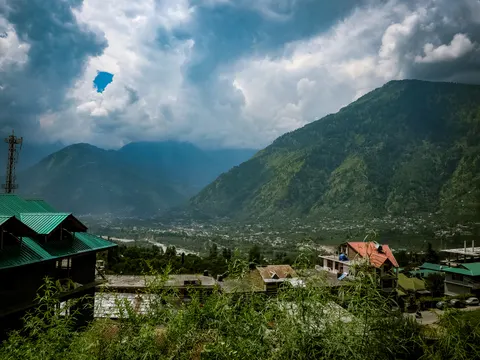 A wide view down a green Himalayan valley, houses with green and red metal roofs in the foreground, a braided river and a spread-out town on the valley floor, forested ridges and heavy cloud above.