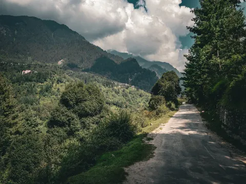A narrow tarmac road curving out of the frame to the right, a stone retaining wall and trees on the right, the wooded valley and a white building on the far slope to the left, cloud above.