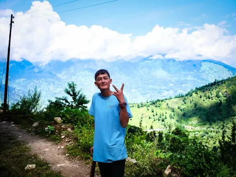 A young man in a pale blue t-shirt standing on the dirt trail to the Chanderkhani pass, smiling and holding up three fingers, a wooden power pole to the left and the Kullu Valley opening out behind him under forested ridges and cloud.