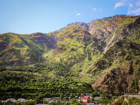 A wide view of a green valley under high grassy ridges, terraced orchards across the middle slope, a red-roofed house and other buildings low on the right and a boulder-strewn river bed at the lower left.