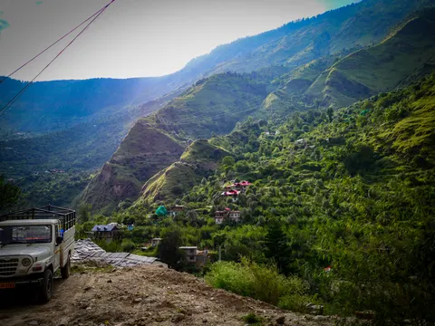 A white pickup truck parked on rough ground at the left with stacked corrugated sheets beside it, a steep green valley beyond, houses and bright pink metal roofs scattered across the far slope and the sun low behind the ridge.