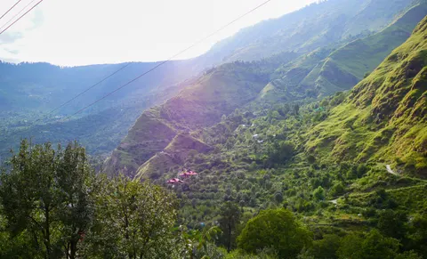 A wide backlit view across a green valley, grassy ridges cut by gullies, houses with bright red roofs on the middle slope, power lines crossing the sky and trees in the lower left corner.