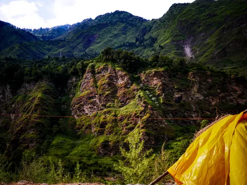 A rocky spur under steep grass with a landslide scar on the slope behind it, an orange rope running across the frame, a yellow tarpaulin at the lower right and hazy ridges beyond.