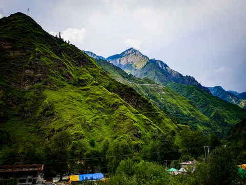 A steep green ridge on the left, a bare rock peak under cloud in the middle distance, power lines crossing the sky and buildings with blue and yellow tarpaulins low on the valley floor.