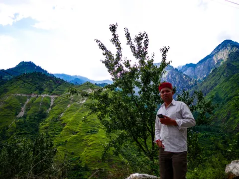 A man standing at the right of the frame in a white checked shirt, a red and gold Himachali cap and dark trousers, holding a phone, a young tree beside him and steep green ridges with a rocky peak behind.
