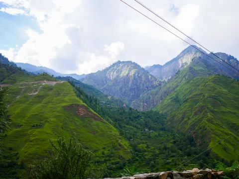A broad valley view, steep grassy ridges cut by pale slide scars, bare rocky peaks in the middle distance, power lines crossing from the upper right and a stone wall at the lower right.