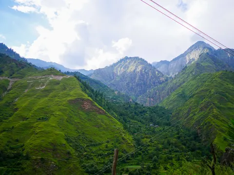The same green ridges and rocky peaks, a wooden fence post in the lower centre, power lines running across the frame and small buildings on the far slope.