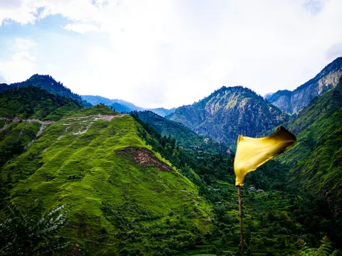 A yellow cloth flag blowing from a thin pole at the right of the frame, steep green ridges scarred by landslides behind it, a rocky peak at the right and a small village on the far slope.