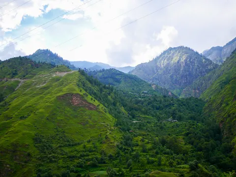 A wide view of a green valley between steep grassy ridges, power lines crossing the top of the frame, a cluster of buildings among trees in the middle distance and small cultivated plots on the floor below.