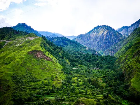 The same green valley framed slightly lower, terraced plots and scattered trees on the floor, a building with a dark roof in the middle distance and bare rock on the ridge behind.