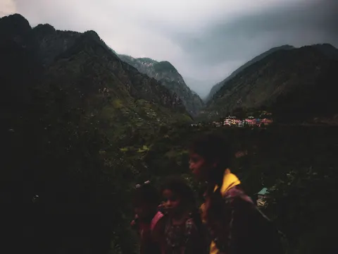 Three children in the foreground at the right seen in profile against a dark valley, a small child in pink at the left, a girl with braids in a patterned top in the centre and an older child in a yellow top and patterned shawl at the right, a brightly painted village on the far slope and cloud filling the head of the valley behind.