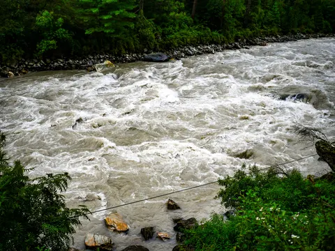A close view straight down onto white rapids filling the frame, boulders breaking the surface, a stone bank and trees along the top edge and a wire crossing the lower part of the frame.