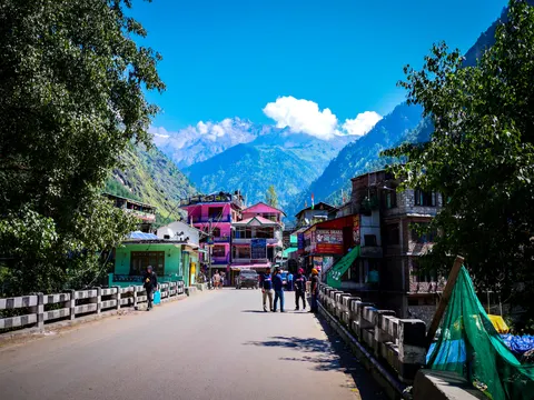 A road running into a small mountain town, painted concrete guesthouses and a dhaba sign either side, people standing in the road and a snow peak showing between forested ridges under a deep blue sky.