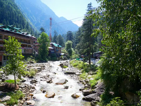 A shallow river running white over boulders through town, hotel blocks with green tin roofs along the left bank, deodars and a red phone mast behind and a wooded ridge above.