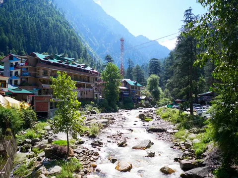 The river running down the middle of the frame, hotels stepping along the left bank, parked cars at the right and forest closing the valley behind.