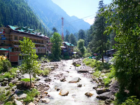 White water over boulders with hotels and shopfronts along the bank, wires crossing the frame and deodars on the slope behind.