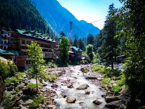 The river at Kasol framed a little wider, buildings at the left, cars parked at the right and the valley narrowing into forest upstream.