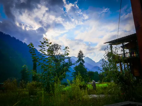 Evening light breaking through cloud over the peaks, tall weeds and young trees across the foreground and the balcony of a wooden guesthouse at the right, with a person standing out in the grass.