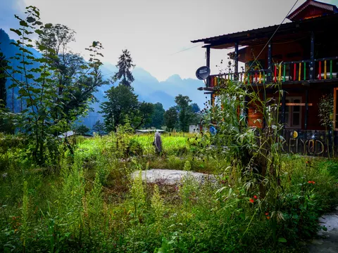 A two storey wooden guesthouse with a painted balcony rail and a satellite dish, lettering painted along the boards, rank grass and flowers in front of it and hills fading into mist behind.