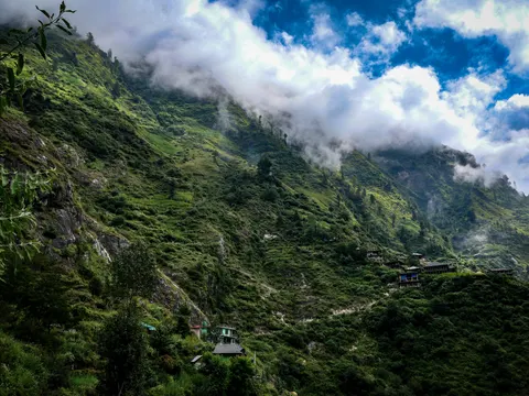 Steep green Himalayan valley with scattered houses below low cloud.