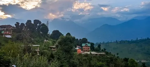 A wide evening view over a ridge settlement, houses with green metal roofs and one orange building set among conifers, layered blue ridges and cloud behind with the sun breaking through.