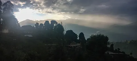 A dark evening panorama, silhouetted conifers and houses on a ridge at the left, shafts of light falling across layered ridges and a misty valley beyond.