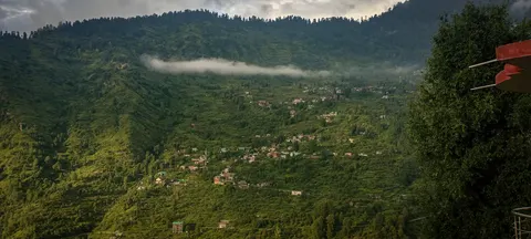 A wide view of a steep green mountainside with houses scattered across the mid-slope, a long band of white cloud lying across it, conifer forest along the ridge top and a tree carrying a red marker at the right edge.
