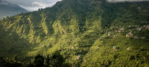 A panorama of a steep green mountainside cut by erosion gullies, scattered houses and a road traversing the slope, conifers along the crest and cloud at the left.