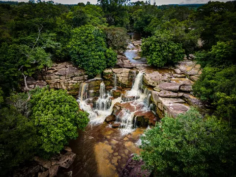 A wide waterfall dropping over layered sandstone ledges into a brown pool, forest crowding both banks, seen from above.