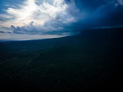 The edge of a forested plateau under a dark storm front, a valley cut into the slope below.