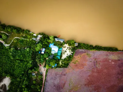A riverbank from above: a house with a blue roof, gardens, and a bare plot of red earth beside the water.