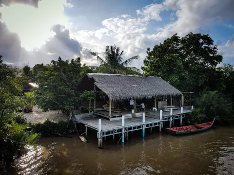 A thatched timber pavilion standing on stilts over a river, a red-painted longboat moored at its deck.