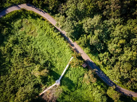 A road curving round a wooded hillside, a cleared green slope inside the bend, seen from above.